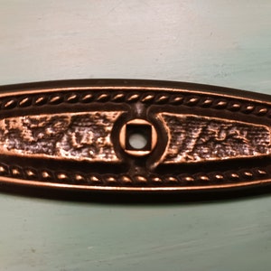 May include: A rectangular, bronze-colored metal plate with a rope-like border and a square hole in the center. The plate has a textured surface.