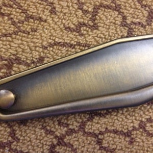 May include: A brushed brass cabinet pull with a rounded rectangular shape and a single round knob. The pull has a slightly curved edge.