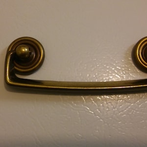 May include: A gold-toned metal cabinet pull with a rounded, decorative design. The pull has a simple, curved shape and is designed for use on cabinets or drawers.