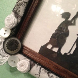 May include: A wooden picture frame with a white lace trim and decorative buttons. The frame contains a silhouette image of two figures, one holding a shovel and the other standing on a platform.