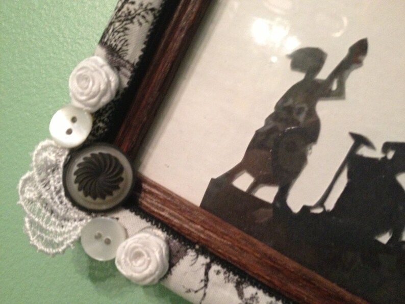 May include: A wooden picture frame with a white lace trim and decorative buttons. The frame contains a silhouette image of two figures, one holding a shovel and the other standing on a platform.