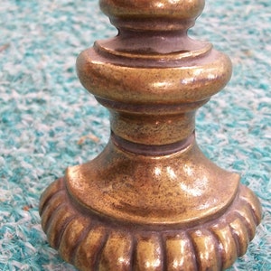 May include: A gold-toned metal finial with a rounded top and a decorative, fluted base. The finial is sitting on a blue and white textured surface.