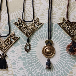 May include: Three gold necklaces with intricate designs and bat-shaped charms. Each necklace has a different style of pendant, including a round pendant, a bat pendant, and a triangular pendant. The necklaces are on a patterned fabric background.