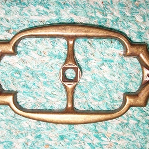 May include: A brass-colored metal decorative plate with a square hole in the center and a decorative design around the edges.
