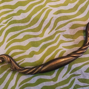 May include: A vintage-style, ornate, brass cabinet pull with a twisted design. The pull is in the shape of a curved handle with decorative details.