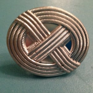 May include: A silver-toned metal ring with a woven design. The ring is oval-shaped and has a smooth, reflective surface.