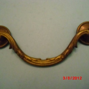 May include: A gold-toned metal drawer pull with a decorative, curved design. The pull has two round ends with a raised center.