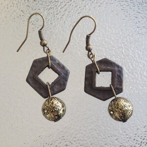 May include: A pair of handcrafted earrings with a geometric design. Each earring features a dark brown, open-center, diamond-shaped pendant suspended from a brass hook. A textured, gold-colored ball hangs below the pendant. The earrings are displayed against a textured, light-colored background.