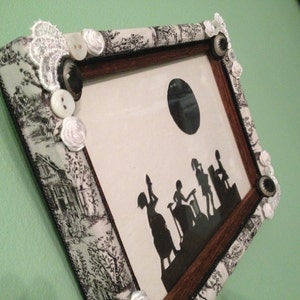 May include: A black and white silhouette of people in a vintage style picture frame. The frame is decorated with white lace, buttons, and flowers.