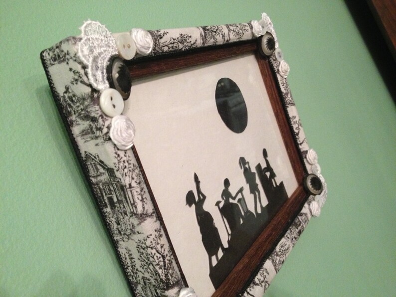 May include: A black and white silhouette of people in a vintage style picture frame. The frame is decorated with white lace, buttons, and flowers.