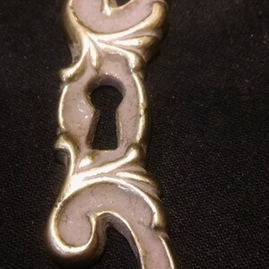 May include: A decorative metal keyhole cover with a pink enamel finish and ornate scrollwork design.
