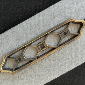 May include: A decorative brass drawer pull with a geometric design. The pull has a hexagonal pattern and two round knobs on each end.