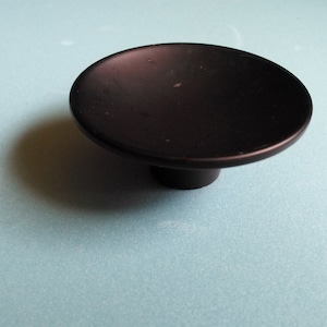 May include: A dark brown, round cabinet knob with a flat top and a short, cylindrical base. The knob is made of metal and sits on a light blue surface. This hardware is suitable for drawers and cabinets.