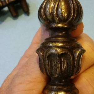 May include: A close-up of a gold-toned metal finial with a decorative, ornate design. The finial has a rounded top and a base with a scalloped edge.