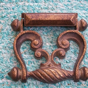 May include: A decorative, ornate, gold-toned metal handle with a twisted design. The handle has a rectangular top and two ornate loops on either side.