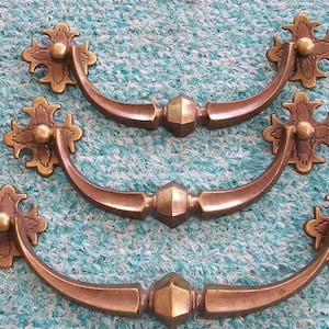 May include: Three antique brass drawer pulls with ornate floral designs and a curved handle. The pulls are arranged in a stack, with the top pull slightly offset from the others.