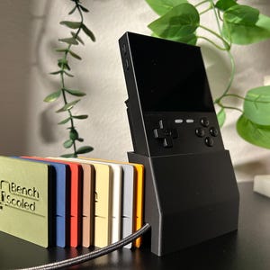 May include: A black handheld gaming console with a screen and buttons. The console is sitting in a black stand with a yellow accent. The console is next to a set of seven colorful cards with the text "Bench Scoled" on the top card.