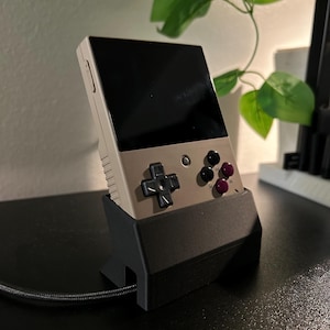 May include: A beige handheld gaming console with a black screen and black buttons is sitting in a black stand. The console is connected to a black cable.