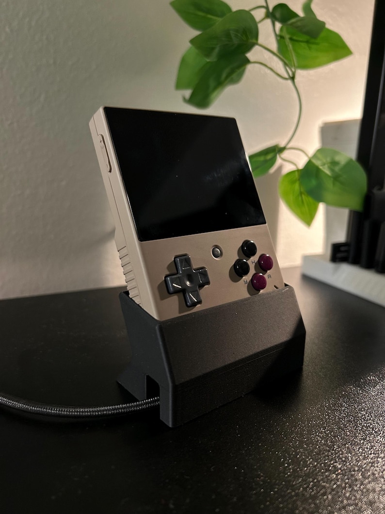Miyoo Mini plus handheld gaming device charging in the black dock, shown on a desk. This Miyoo is charging with an Anker cable (not included).