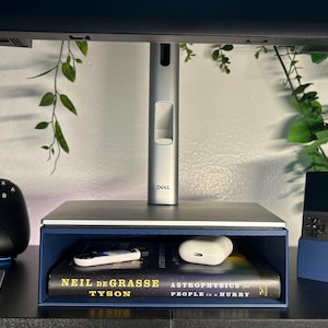 May include: A blue desk organizer with a monitor stand, a book titled "Astrophysics for People in a Hurry" by Neil deGrasse Tyson, a white wireless earbud case, and a black and blue handheld gaming device.