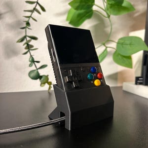 May include: A black handheld gaming console with a screen, buttons, and a directional pad. The console is sitting in a black stand on a dark surface.