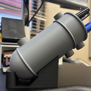 May include: A gray 3D printed pen holder with a cylindrical shape and multiple holes for pens. The holder is angled on a black stand.