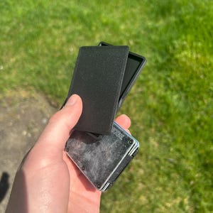 May include: Black wallet with a textured cover, held open to reveal the interior. The wallet has a metal frame and a contrasting black interior. The background is a blurred green lawn.