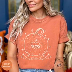 May include: Burnt orange t-shirt featuring a cream-colored pumpkin design with a heart-shaped flame and floral accents. The shirt has short sleeves and a relaxed fit, perfect for fall. The text "Burnt Orange" is visible.