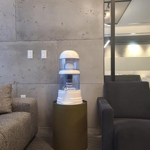 May include: A white water filtration system with a clear filter and a brown pedestal base. The system is in a room with a concrete wall and a gray couch.