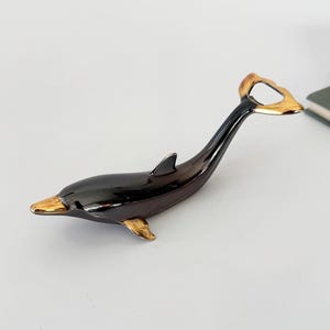 Puede incluir: Un abrebotellas con forma de delfín negro con detalles dorados en la nariz, las aletas y el abridor. El abridor es de metal y tiene un acabado brillante. El delfín está sobre una superficie blanca.