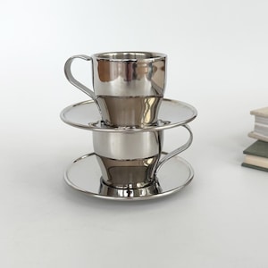 Könnte beinhalten: Zwei gestapelte Espressotassen mit passenden Untertassen aus poliertem, silberfarbenem Metall. Jede Tasse verfügt über einen geschwungenen Henkel und eine glatte, reflektierende Oberfläche. Die Tassen sind übereinander angeordnet.