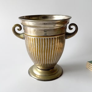 Puede incluir: Un recipiente metálico en forma de urna con borde y base en tono plateado pulido, que contrasta con un cuerpo de costillas verticales en un acabado bronce dorado cálido. La pieza incluye dos asas simétricas curvadas en los lados.
