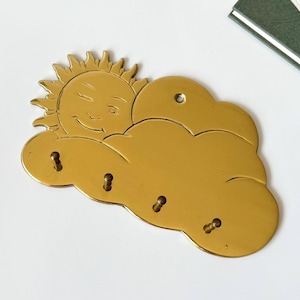 May include: A gold-colored key holder shaped like a cloud with a smiling sun peeking out. The key holder has four key hooks and a hole for hanging. The design is whimsical and decorative.