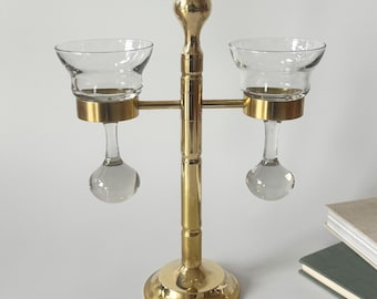 May include: A brass candle holder with two clear glass cups. The candle holder has a round base, a central stem, and two horizontal arms holding the cups. The cups have a bulbous base and a flared rim. The overall design is symmetrical.