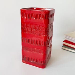 May include: A bright red ceramic vase, rectangular in shape, with a glossy finish. The vase has a textured design with horizontal bands and embossed patterns. It sits on a white surface, with a stack of books visible in the background.