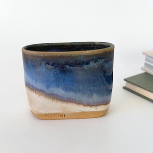 May include: A handmade ceramic tumbler with a unique blue and white glaze. The top rim is dark blue, transitioning to lighter blues and white at the base. The word "KAMINI" is stamped on the bottom. A minimalist design for home decor.