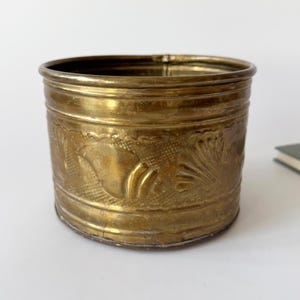 Könnte beinhalten: Ein Vintage-Messingbehälter mit geätzten Blumen- und Fischmotiven. Das zylindrische Gefäß hat eine leicht angelaufene, goldene Oberfläche. Der Rand und der Boden sind klar definiert, und der Körper weist detaillierte Reliefmuster auf. Ein dekoratives Stück.