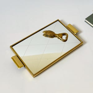 Op de afbeelding: Een rechthoekig spiegeldienblad met een goudkleurig metalen frame en twee bijpassende handgrepen aan de zijkanten. Een messingkleurige flesopener in de vorm van een vis ligt op het reflecterende oppervlak met een subtiel ruitpatroon.
