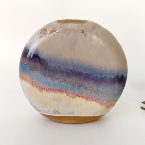 Könnte beinhalten: Eine kugelförmige Keramikvase mit einer strukturierten, mehrfarbigen Glasur. Die Farben umfassen Beige, Blau, Lila und einen Hauch von Rot. Die Vase steht auf einem hellbraunen Sockel und hat eine kleine Öffnung oben.