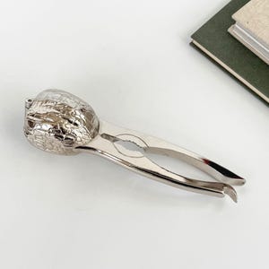 Könnte beinhalten: Ein silberfarbener Nussknacker aus Metall mit einem strukturierten, walnussförmigen Kopf und zwei länglichen, glatten Griffen. Das Werkzeug verfügt über ein Scharnierdesign mit einem gezackten inneren Greifbereich zum Knacken von Schalen.