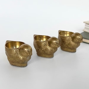 Könnte beinhalten: Drei Eierbecher aus Messing in Form kleiner Vögel. Jeder Becher verfügt über eine strukturierte Metalloberfläche mit detaillierten Flügel- und Kopfgravuren, eine hohle Mitte zum Halten eines Eies und einen flachen Boden für Stabilität.