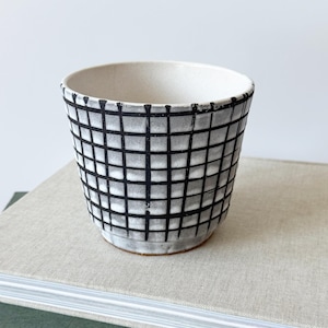 Puede incluir: Una pequeña taza de cerámica hecha a mano con un patrón de cuadrícula. La taza es blanca con un diseño de cuadrícula negra y una base natural sin esmaltar. La taza está sobre una pila de libros.