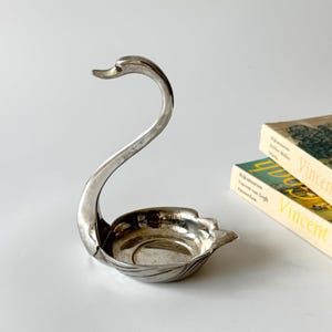Könnte beinhalten: Eine silberfarbene Metallschale in Schwanenform mit einem langen, geschwungenen Hals und einer abgerundeten, flachen Schalenbasis mit dezenten flügelartigen Details. Das Objekt hat eine polierte, reflektierende Oberfläche und dient als kleiner Behälter.