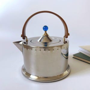 May include: A polished stainless steel teapot with a cylindrical body, a short spout, and a conical lid topped with a small blue spherical knob. The teapot features a curved wooden handle attached to the sides with metallic hardware.