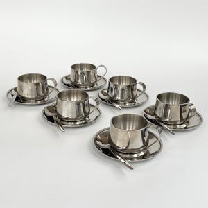 May include: A set of six polished stainless steel cups, each resting on a matching circular saucer with a small spoon placed alongside. The cups feature a simple cylindrical shape with a curved handle, reflecting light on their metallic surface.