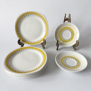 May include: A collection of white ceramic plates and saucers featuring a yellow and blue circular pattern. The plates have a square shape with rounded edges. The saucers and plates are displayed on brass stands, showcasing a vintage tableware set.