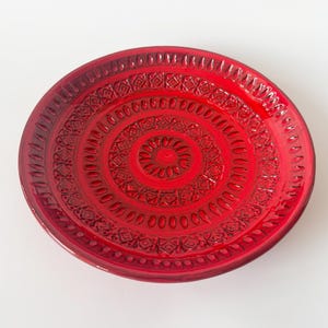 Op de afbeelding: Een ronde, rode keramische schaal met een gedetailleerd, reliëfpatroon. De schaal heeft concentrische cirkels en geometrische ontwerpen, met een glanzende afwerking. De rand heeft kleine, gelijkmatig verdeelde inkepingen. De schaal is een levendige rode kleur.