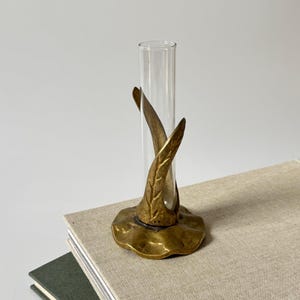 Könnte beinhalten: Eine Vase aus einem klaren Glas-Reagenzglas, gehalten von einem messingfarbenen Metallsockel. Der Sockel zeigt ein strukturiertes, organisches Blattdesign, das den Boden des Glasrohrs umschließt und auf einer gewellten Metallplattform ruht.