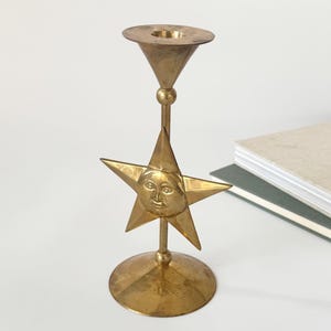 Könnte beinhalten: Ein Messing-Kerzenhalter mit Stern-Design. Der Stern hat ein Gesicht in der Mitte und wird von einem schlanken Stiel und einem kreisförmigen Sockel getragen. Die Oberseite des Kerzenhalters hat die Form eines kleinen, umgekehrten Kegels.