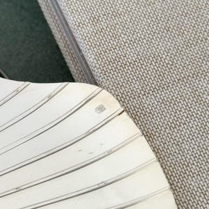 May include: A silver-colored, fan-shaped object with a ridged design. The object has a small emblem in the upper right corner. The background includes a textured, light-colored fabric and a dark green surface.